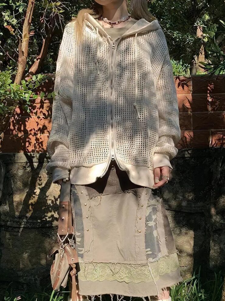 Une personne porte une jupe kaki, un sac et un cardigan à capuche zippé beige. Le cardigan est ouvert en haut et en bas