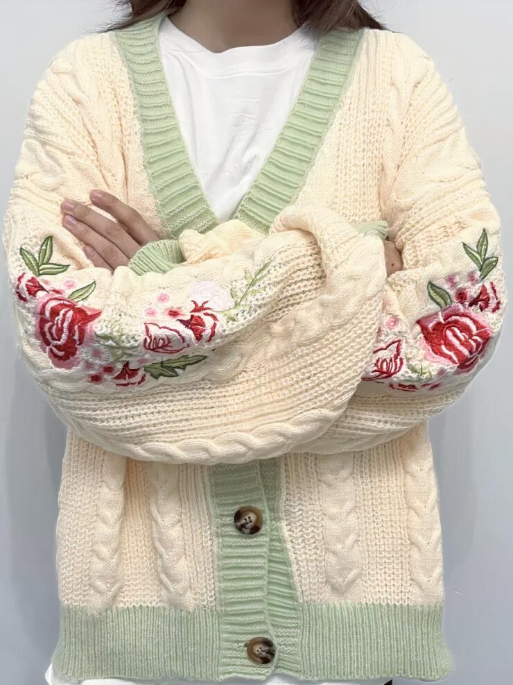 Une personne porte un tee shirt blanc et un cardigan beige et vert avec des fleurs et des torsades.La personne croise les bras