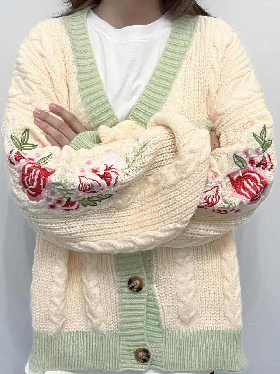 Une personne porte un tee shirt blanc et un cardigan beige et vert avec des fleurs et des torsades.La personne croise les bras