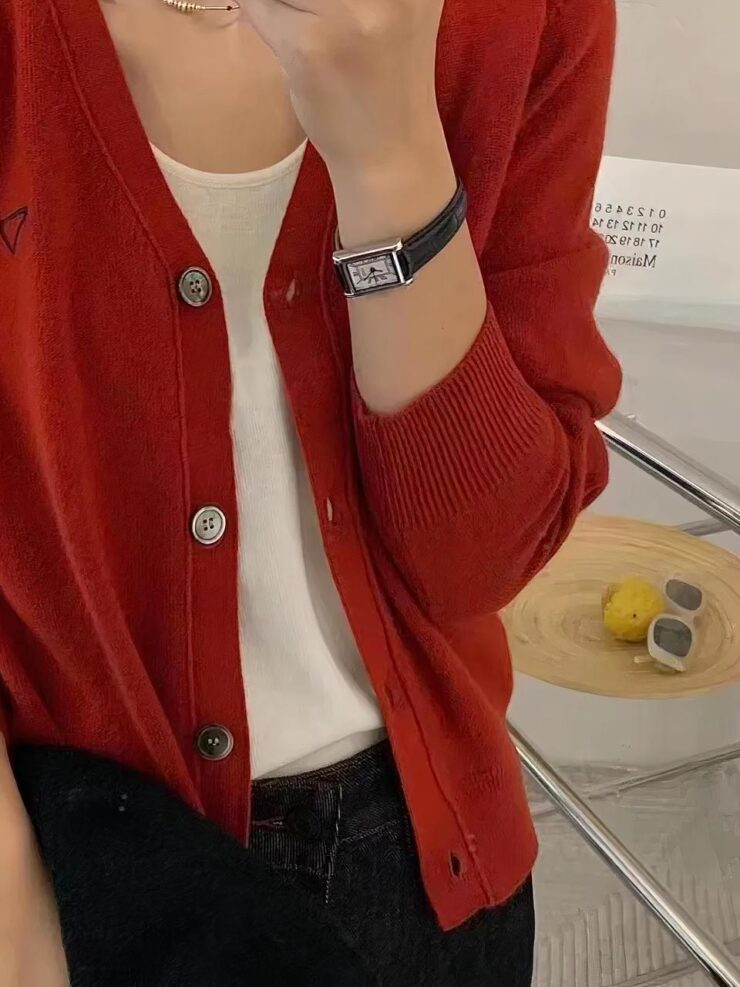 Photo d'une jeune femme portant un cardigan rouge en cachemire col V à quatre boutons, ouvert sur un teeshirt blanc et un pantalon noir, dans un décor beige