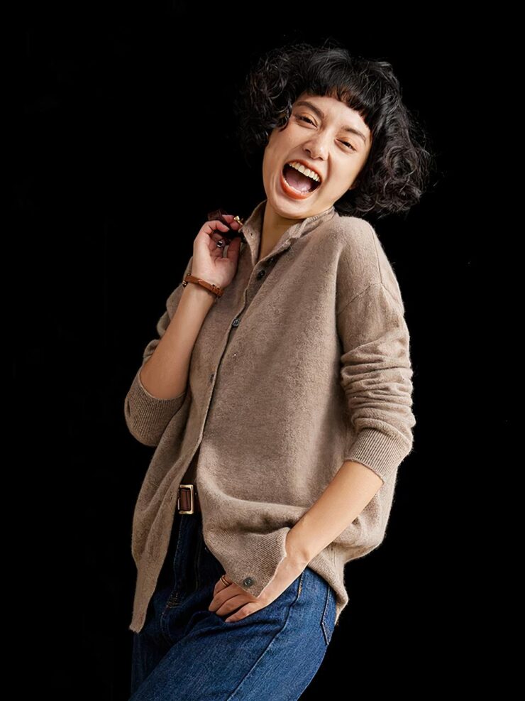 Photo d'une jeune femme debout de trois quart profil, penchée légèrement en arrière et riant avec la main droite sur la bandoulière de son sac et la gauche dans la poche de son jean, elle porte un cardigan mi-long en cachemire marron à col montant, le tout sur un fond noir