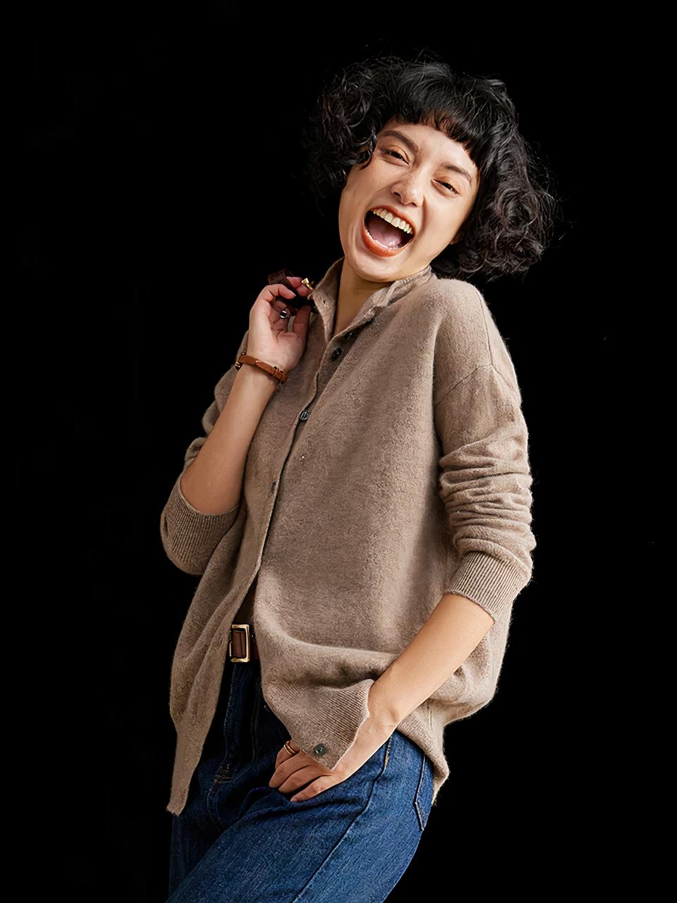 Photo d'une jeune femme debout de trois quart profil, penchée légèrement en arrière et riant avec la main droite sur la bandoulière de son sac et la gauche dans la poche de son jean, elle porte un cardigan mi-long en cachemire marron à col montant, le tout sur un fond noir
