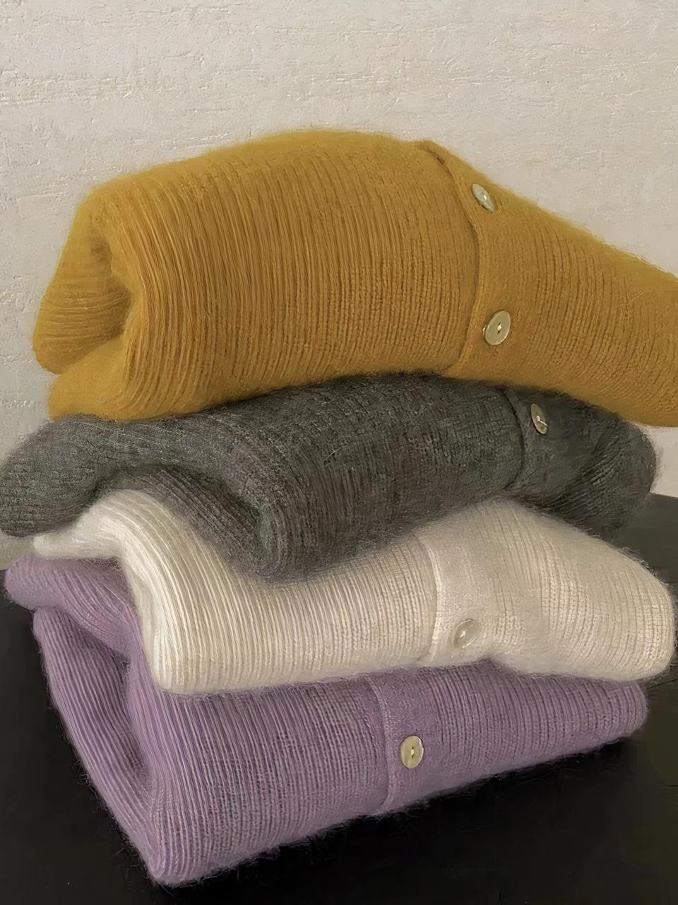 Cardigan cachemire femme fluide Photo représentant les quatre cardigans en cachemire fluides de couleur différente pour femme pliés sur un meuble marron foncé devant un mur beige