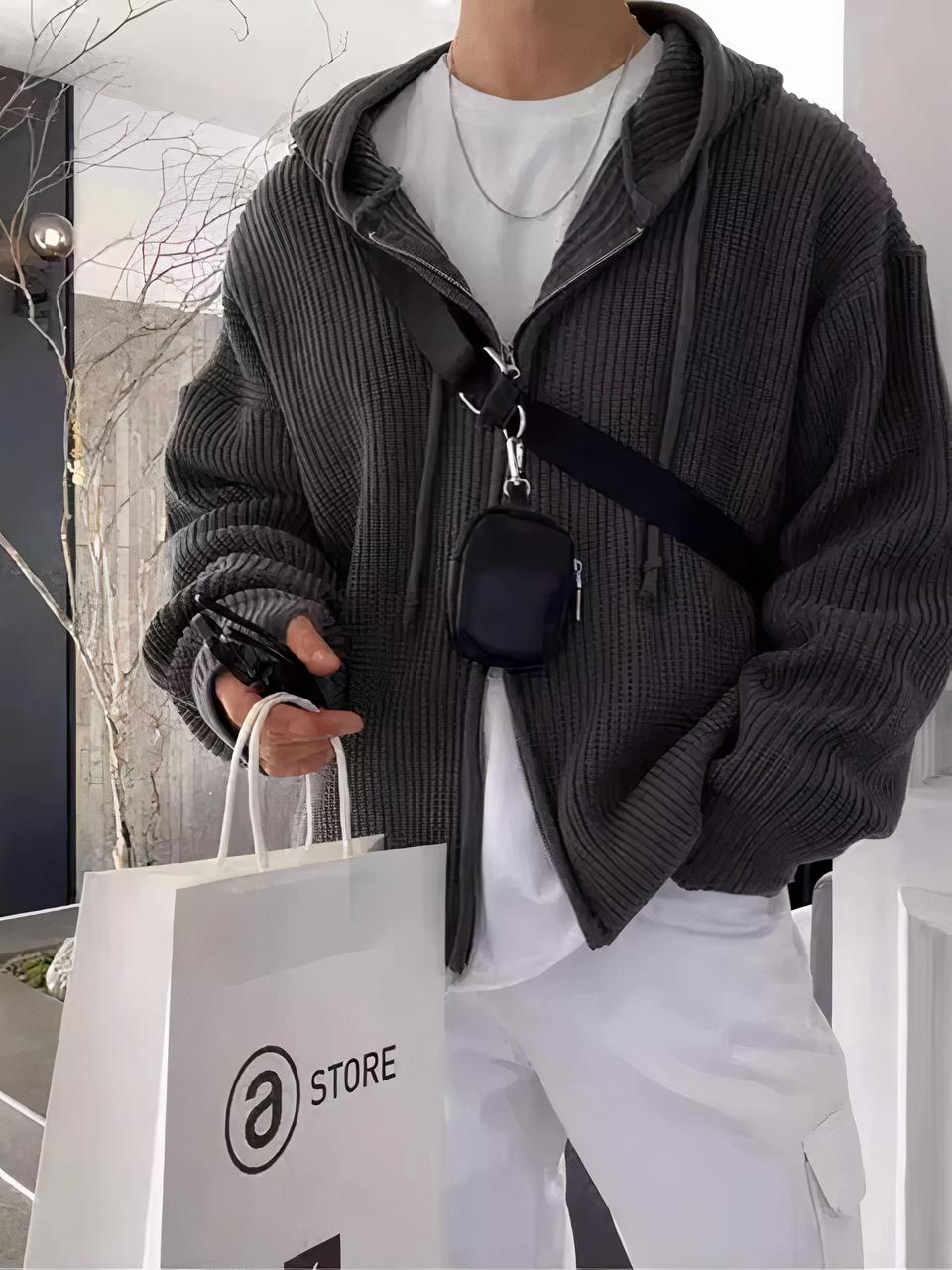 Photo d'un jeune homme debout un sac blanc en papier à la main droite , la main gauche dans la poche de son pantalon blanc large, portant un cardigan gris zippé à capuche sur un teeshirt blanc, le tout dans une pièce à dominance blanche