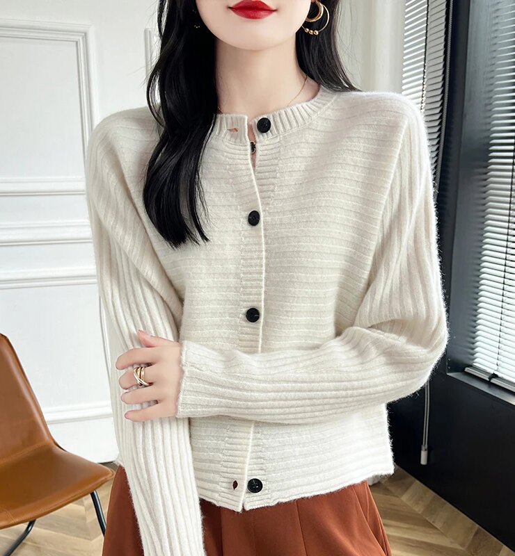 Photo d'une jeune femme debout dans une pièce à parquet en bois et store gris sur baie vitrée avec une chaise marron au second plan, elle porte un cardigan blanc boutonné à col rond, manches longues en laine tricotée en mailles horizontales sur un pantalon large couleur rouille