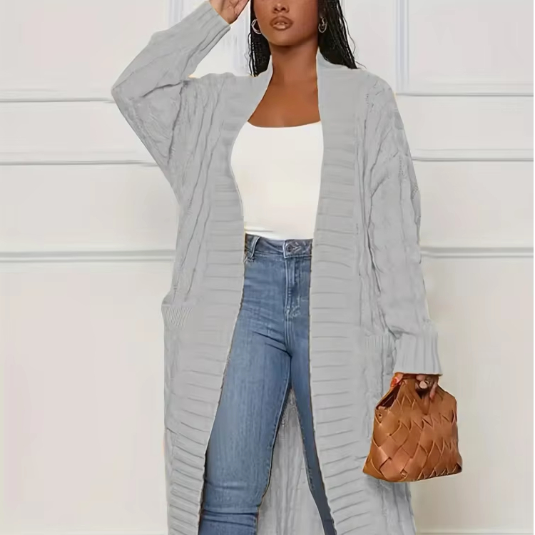 Une femme porte un jean, un top blanc et un gilet long gris clair. Elle porte à la main un sac à main marron