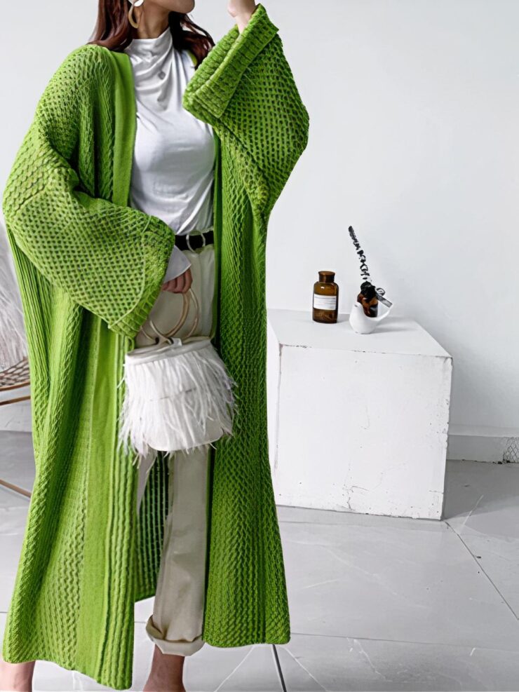 Une femme porte un cardigan long vert, un jean et un haut blanc. Elle a un sac blanc et est prise en photo devant un fauteil et une table blanche