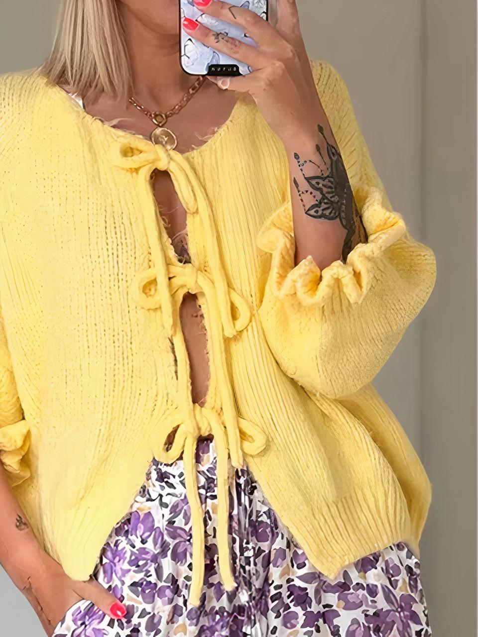 Photo d'une femme debout légèrement penchée sur la droite avec la main dans la poche de son pantalon d'été violet à fleurs et portant un cardigan en maille jaune à nouer devant et à manches 3/4 à volant, la main gauche tenant un téléphone devant le visage, le tout dans une pièce beige