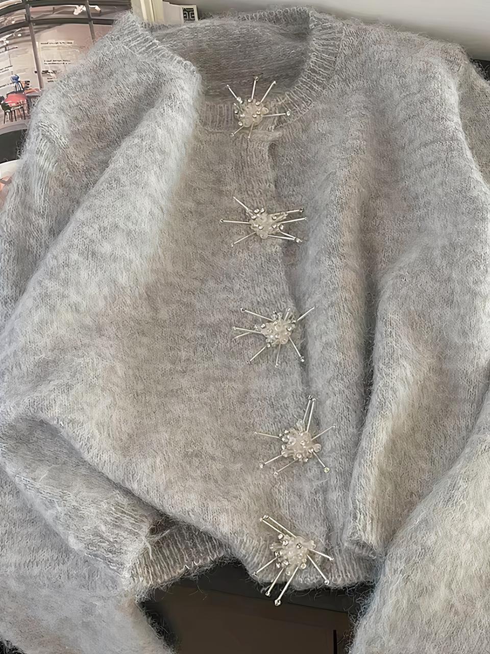 Photo d'un cardigan gris à mailles souples col rond et boutons en forme de flocons à strass, posé sur un meuble marron, un magazine sur la gauche