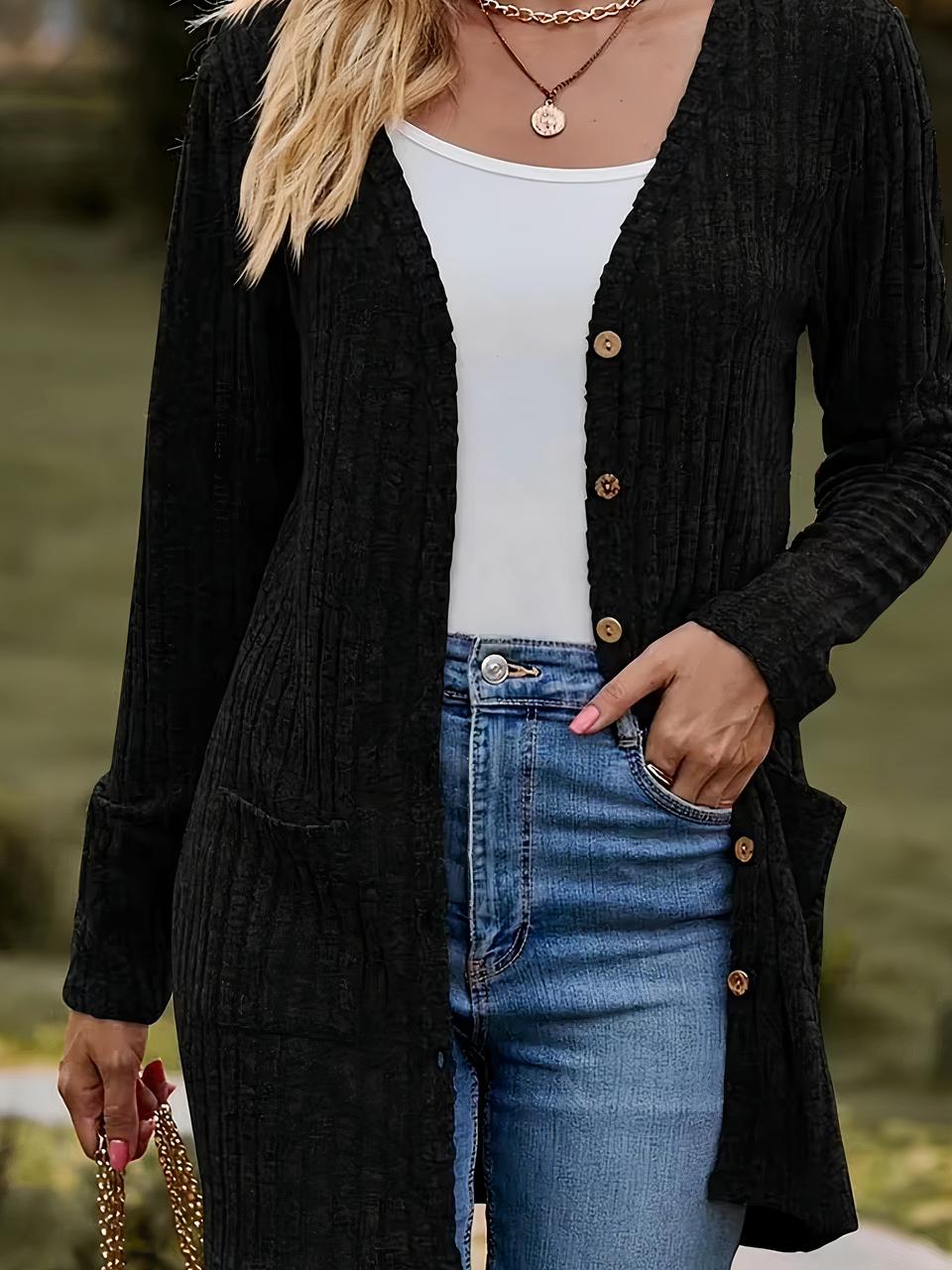 Photo d'une jeune femme debout dans la nature portant un cardigan noir mi-long col V à petits boutons ouvert sur un jean bleu ceinturé et un teeshirt blanc col bateau, ma main gauche dans la poche revolver du jean