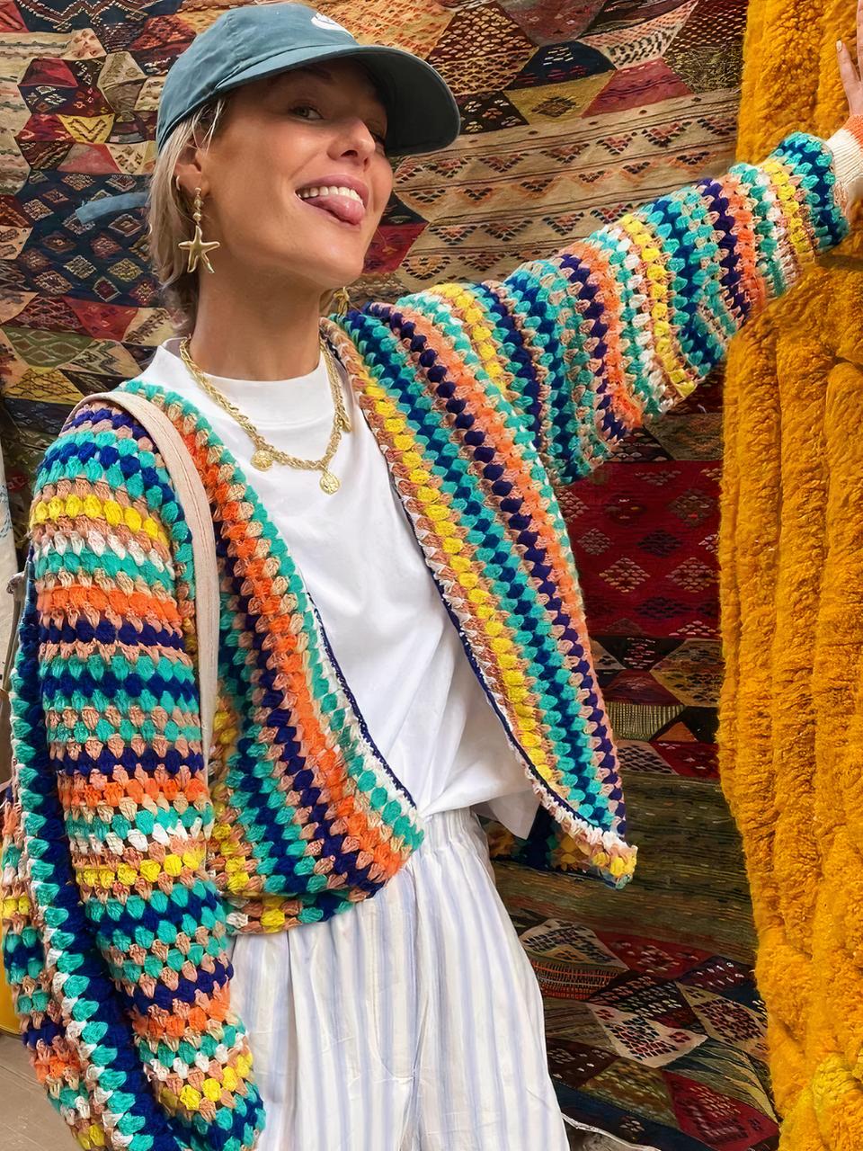 Cardigan rayé élégant pour femme Cardigan à rayures multicolores porté par une femme. Elle porte une casquette bleu un pantalon rayé blanc et beige et un tee shirt blanc. Elle est entouré de tapis de couleurs