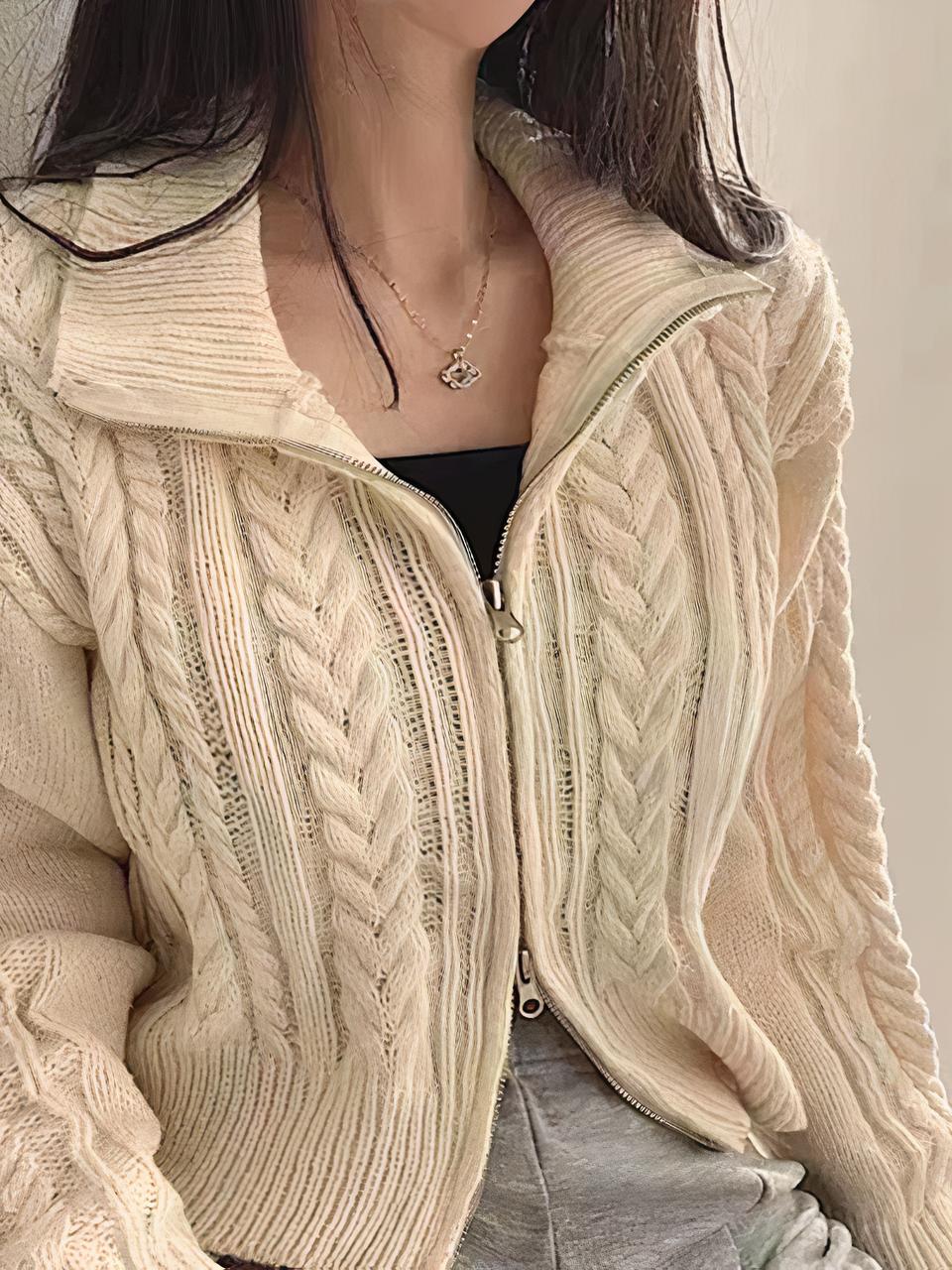 Une femme brune porte un cardigan beige avec des torsades et une fermeture éclair. La fermeture est ouverte en haut et en bas. Elle porte un top noir et un pantalon gris