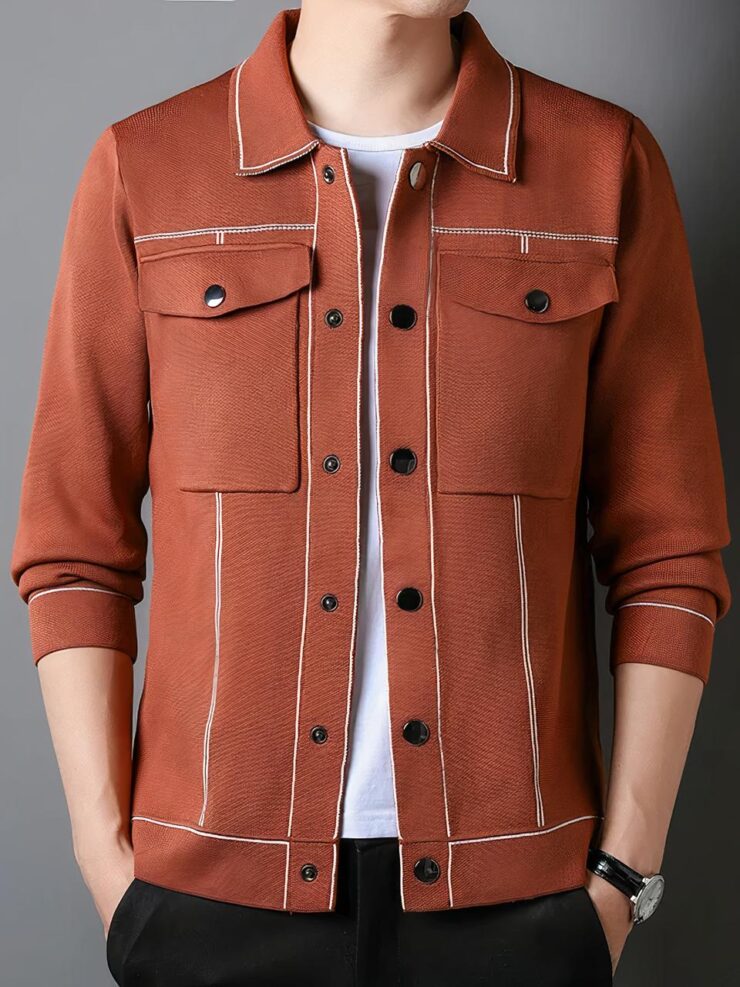 Photo d'un homme debout les mains dans les poches de son pantalon noir et vêtu d'une veste cardigan orange à poches plaquées hautes fermées par bouton pression, six autres boutons pression devant et surpiqures en couleur contrastée sur un teeshirt blanc col rond, le fond est gris foncé à droite et clair à gauche