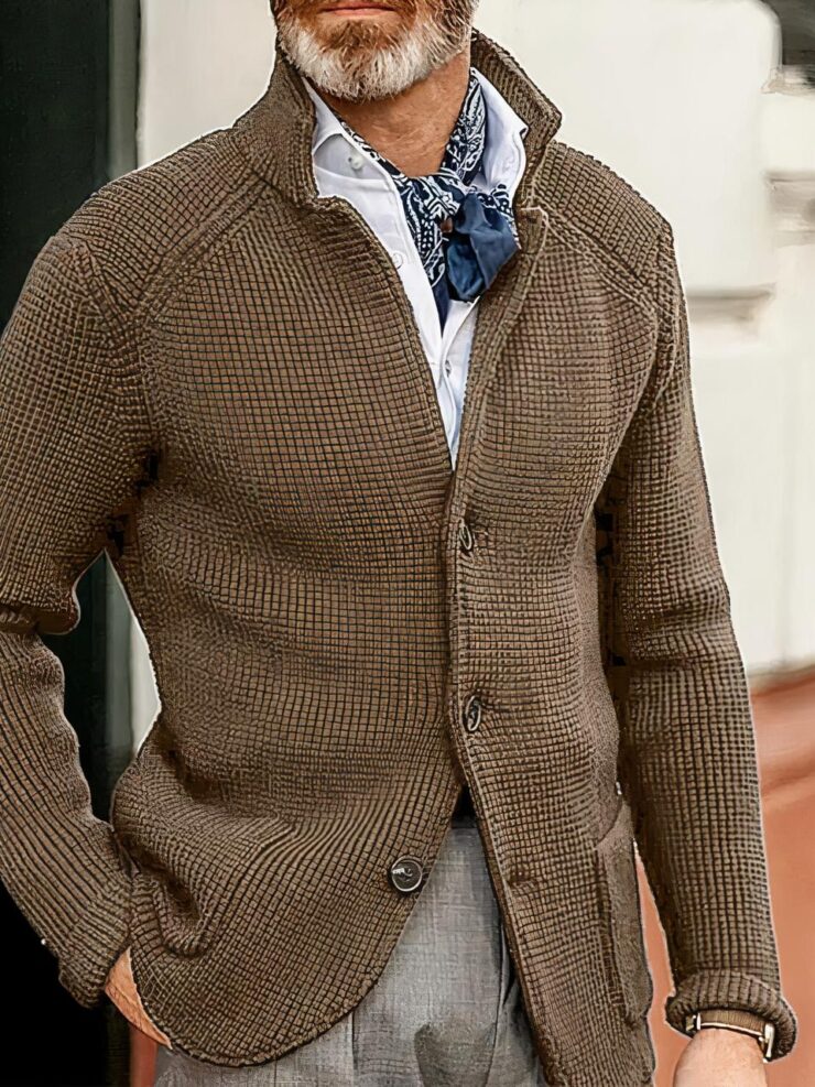Photo d'un homme debout devant un mur d'habitation bicolore, la main droite dans la poche de son pantalon et portant une veste cardigan marron tricotée, fermée par deux boutons sur trois, à col relevé sur une chemise blanche et un foulard bleu autour du cou