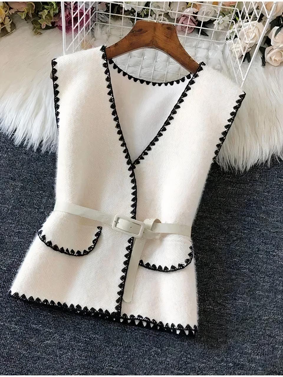 Photo d'un cardigan chic original blanc sans manche blanc encolure en V à contours et bords de revers poche noir, ceinture blanche, sur un cintre posé sur un sol gris avec un morceau de fourrure blanche en haut et bouts de chaussures blanches à gauche