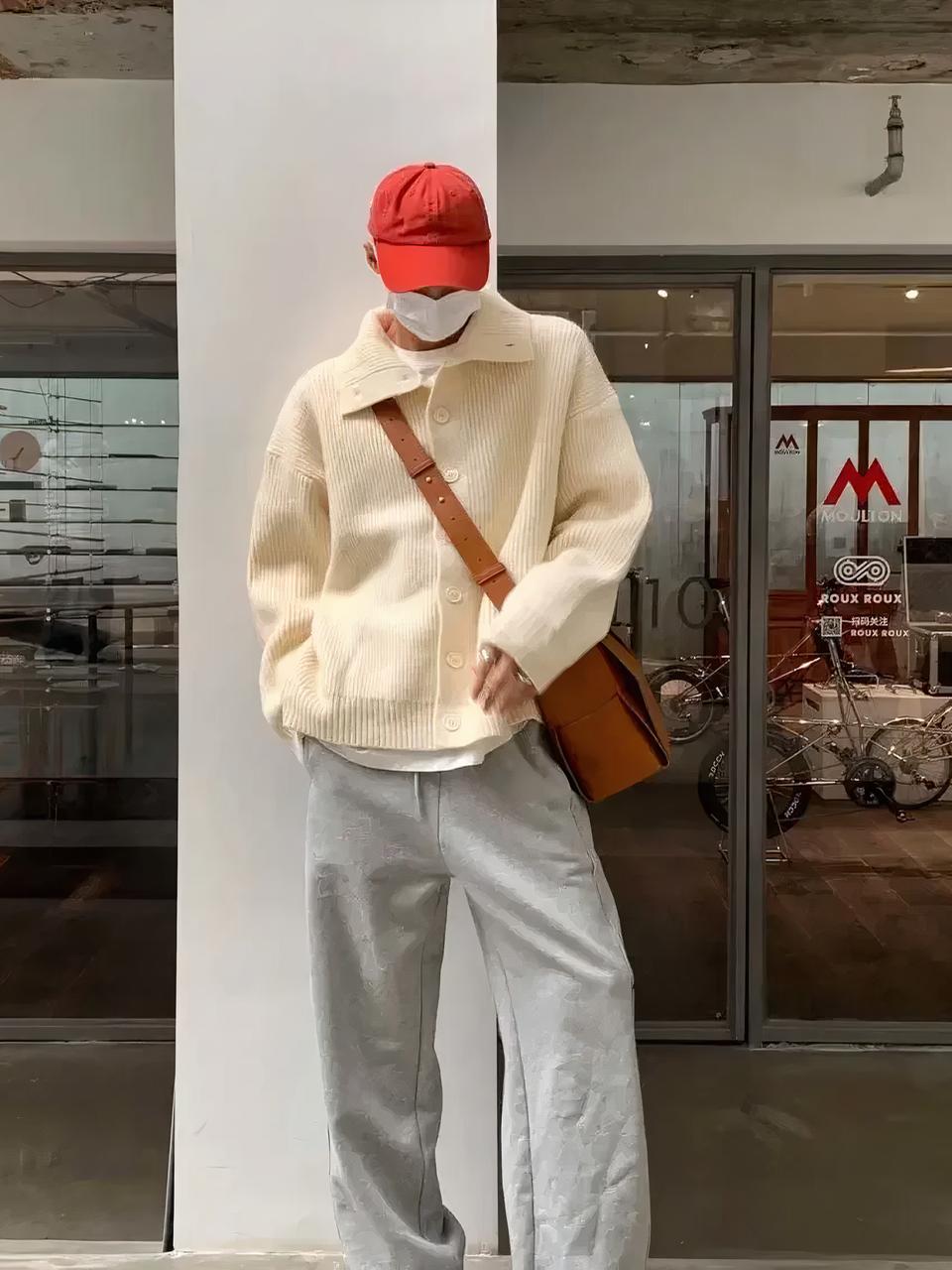 Un homme est adossé à un poteau blanc. Il porte un pantalon gris, un gilet beige large avec un grand col et une casquette rouge. Il a un sac marron en bandoulière.