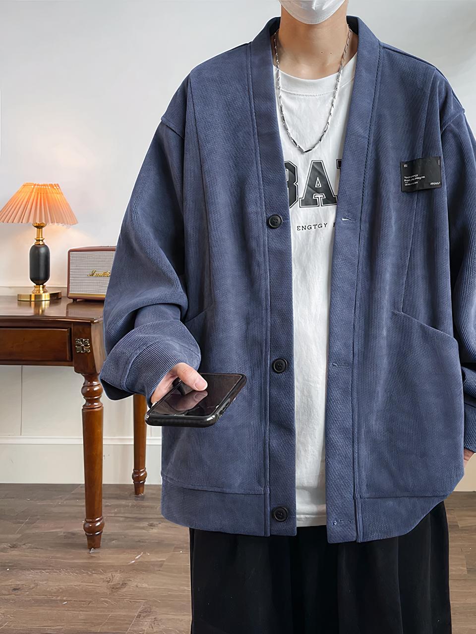 Un homme se trouve dans une pièce et porte un pantalon noir, un tee shirt blanc et une veste bleue longue. L'homme a son portable dans la main