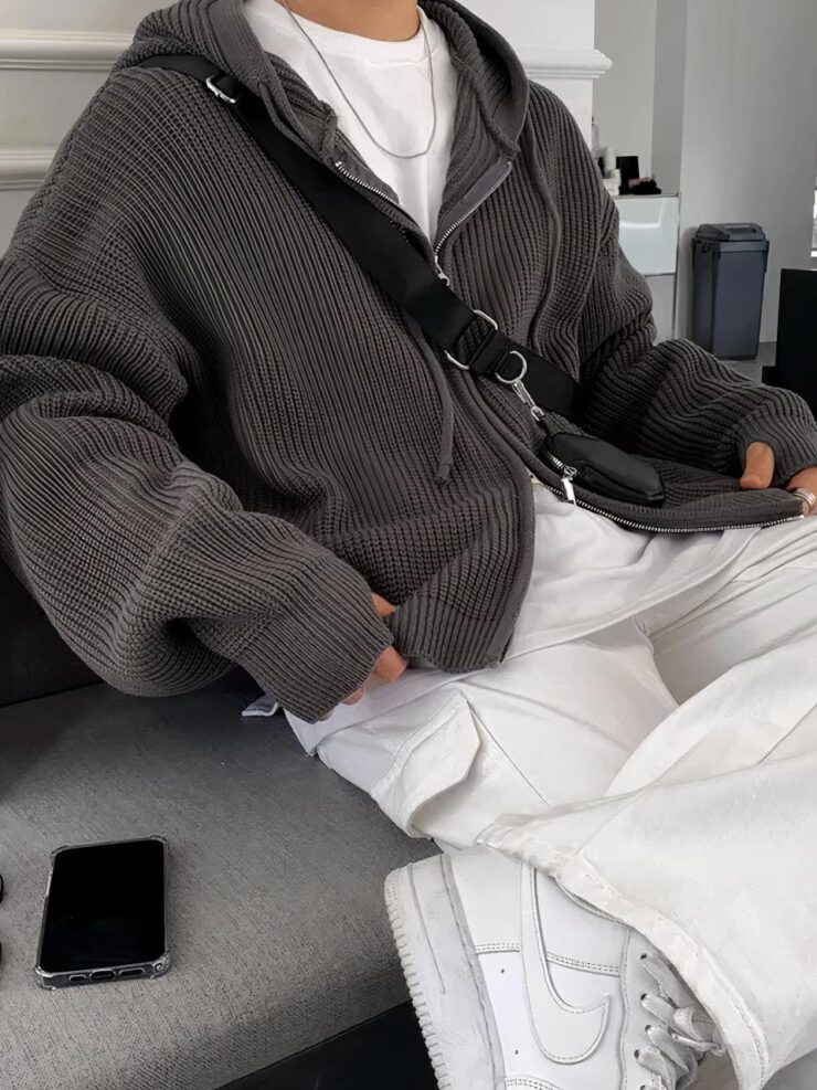 Une homme assis porte un pantalon blanc, des baskets blanches, un tee shirt blanc et un gilet large zippé à moitié ouvert et une capuche