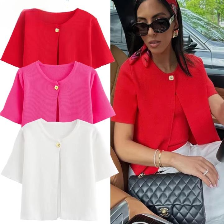 Une femme brune porte tes lunettes de soleil, un cardigan rouge fermé par un bouton doré. Elles est assisse dans une voiture et a un sac à main noir. 1 cardigan blanc, un rose et un rouge sont exposés à côté de la femme